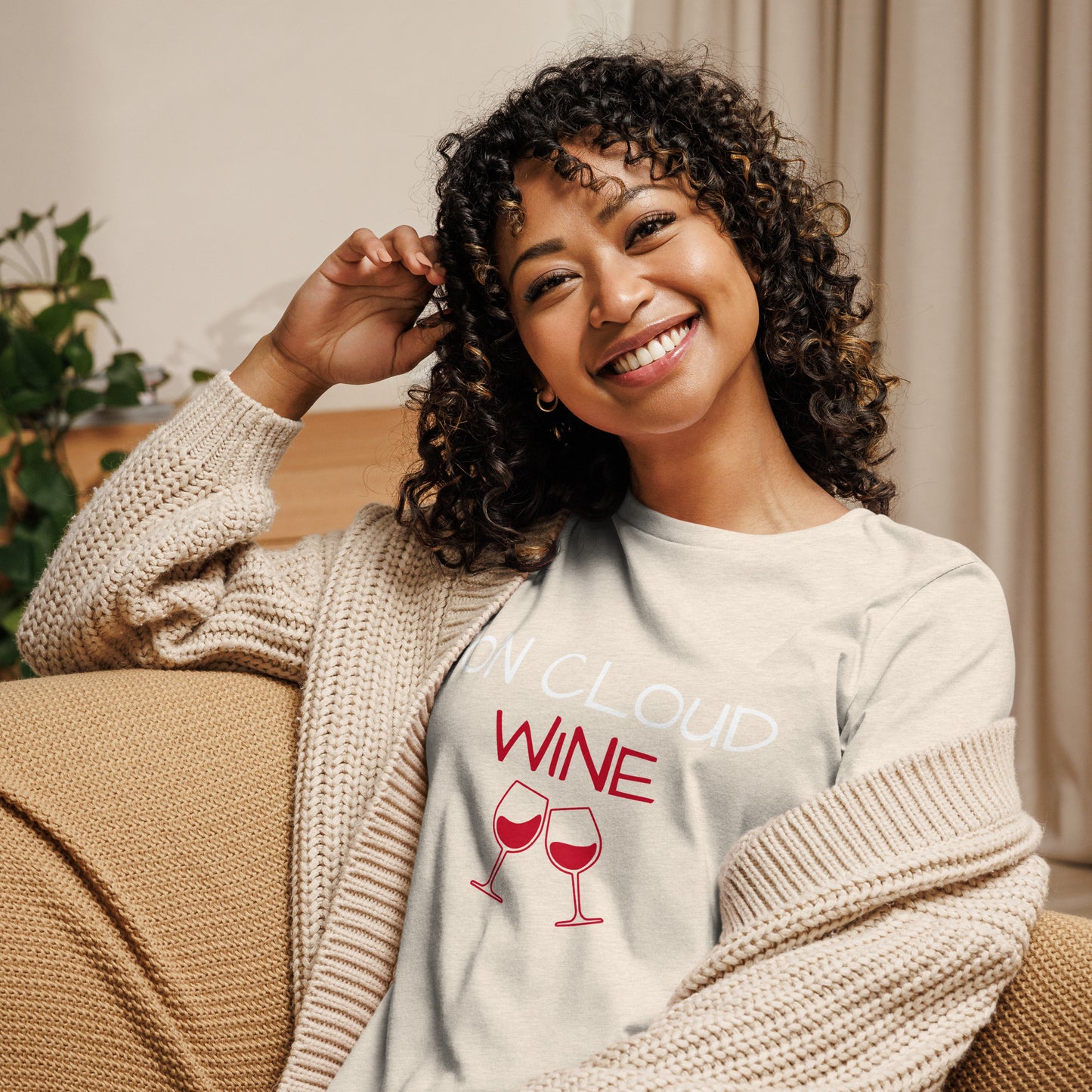 Woman wearing Women's Relaxed T-Shirt with "On Cloud Wine" text and wine glasses graphic, smiling on a cozy sofa.